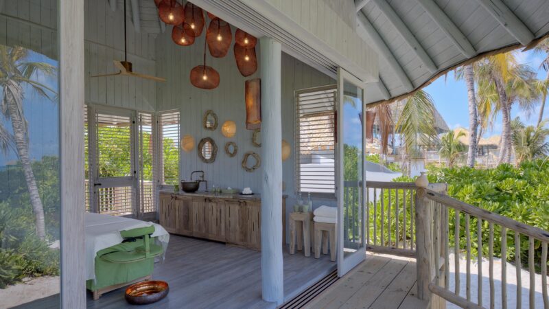 Open-air wellness spa room with a massage bed draped in green and white linens, wooden decor on the walls, a basin, and a tray with spa essentials. Overhead, wicker pendant lights hang from the ceiling. The room opens to a lush tropical garden and is covered by a rustic roof. Experience Maldive Spa serenity here.