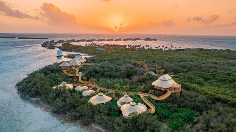 A scenic aerial view of an island resort during sunset. The resort, featuring several thatched-roof huts amidst lush greenery and connected by wooden walkways, glows under the sun's golden hue. Discover Soneva Exclusive Offers as the sun sets over the ocean, casting its warm light on the tranquil waters.