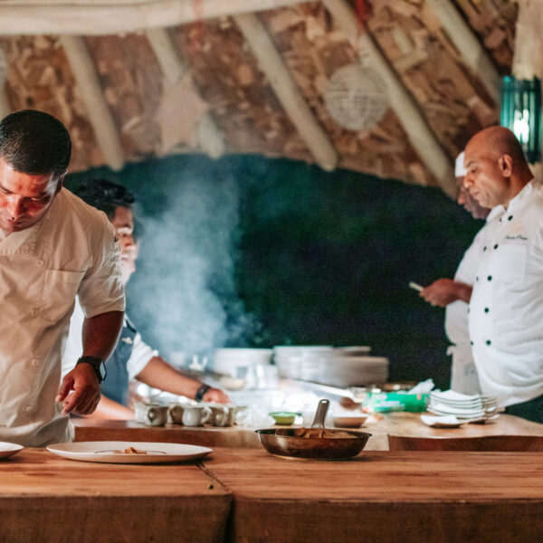 In the rustic, open-air kitchen, chefs in white uniforms diligently craft culinary masterpieces. One chef uses a yellow squeeze bottle to garnish a dish under soft lighting and steam. As plates and utensils clatter gently, the ambiance whispers of Soneva Exclusive Offers enriching this unique experience.