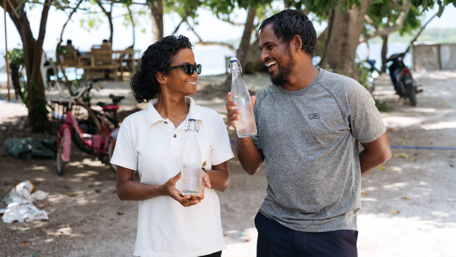 Two people standing outdoors, both smiling and holding glass bottles. The person on the left wears a white shirt and sunglasses, while the person on the right has short hair and wears a gray shirt. There are trees and bicycles in the background, along with a wooden bench that was donated by the Maldives Foundation.