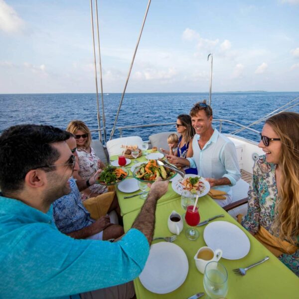 A group of people are sitting around a table on a luxury yacht in the Maldives, enjoying a meal together. The table is set with plates, glasses, and several dishes. The ocean is visible in the background, and the sky is partly cloudy. Everyone appears to be smiling and engaged in conversation.