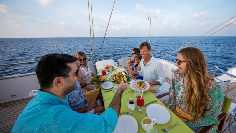 A group of people are sitting around a table on a luxury yacht in the Maldives, enjoying a meal together. The table is set with plates, glasses, and several dishes. The ocean is visible in the background, and the sky is partly cloudy. Everyone appears to be smiling and engaged in conversation.