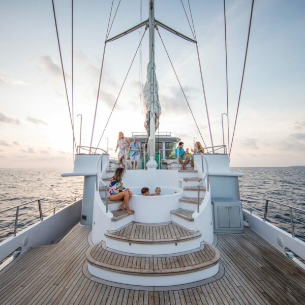 A group of people relaxing on the deck of a luxurious sailboat, anchored at sea during sunset. The spacious deck with curved steps leading up to the mast complements the serene backdrop of calm ocean and scattered clouds, making for an idyllic Maldives luxury yacht holiday.