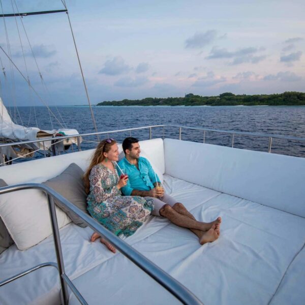 A man and woman relax on a large cushioned bench aboard a luxury yacht during their Maldives holiday, gazing at the ocean under a pastel-colored sky at sunset. The woman holds a drink while they both sit barefoot, enjoying the tranquil scene.