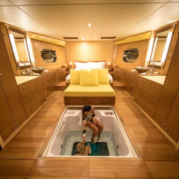 A luxurious Maldives luxury yacht holiday cabin features a large bed with yellow pillows and a modern, wooden interior. At the center, a woman relaxes in a sunken hot tub. The cabin is well-lit, with mirrors and windows on both sides.