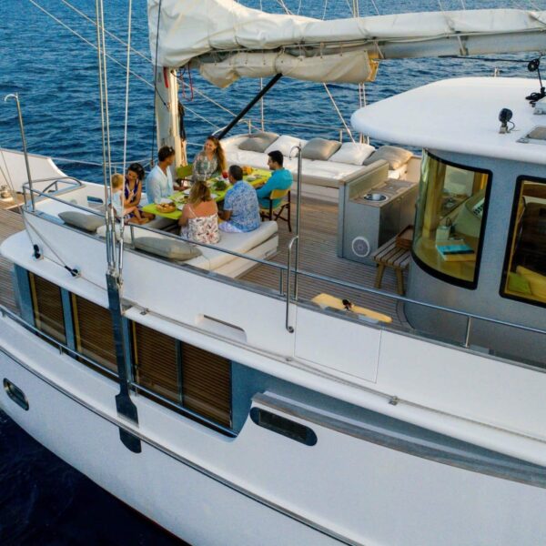 A group of people enjoying a meal on a luxurious white yacht during their Maldives Luxury Yacht Holiday. The yacht sails on calm blue waters, its spacious deck featuring wooden flooring. The sun shines brightly as various dining items and colorful decorations adorn the table.