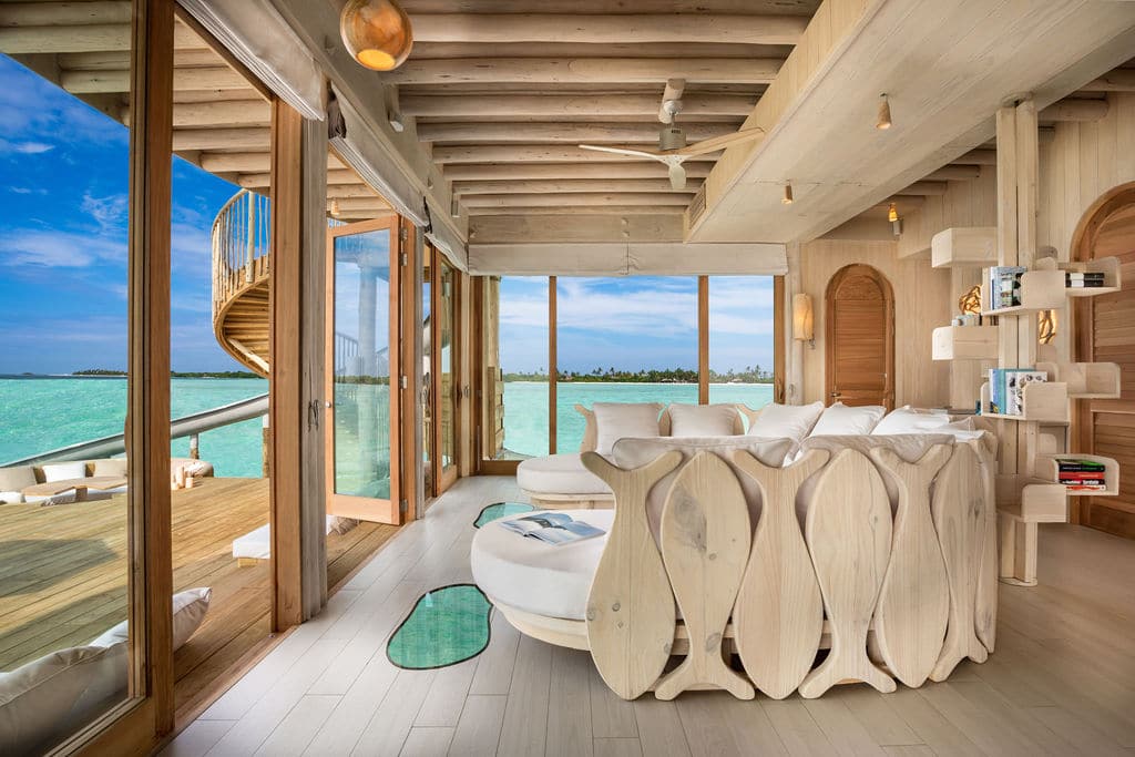 A luxurious, sunlit room at the Maldives Luxury Resort features wooden furnishings and large glass windows offering a stunning view of turquoise waters. The space is decorated with white sofas, unique wooden decor, a ceiling fan, and a spiral staircase leading to an outdoor deck at Soneva Villas.