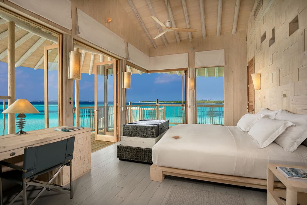 A serene overwater bungalow at the Maldives Luxury Resort, Soneva Villas, with large windows offering a panoramic view of turquoise waters. The airy room has a wooden interior, a cozy bed, a desk, and a seating area. A door leads to a private deck over the water. Natural light fills the space.