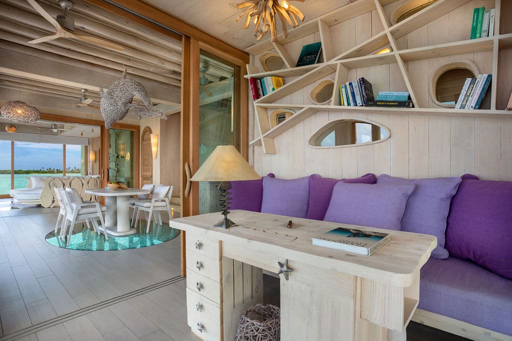 A cozy living area with a wooden desk and purple cushions on a bench. A unique bookshelf with various books is mounted on the wall above the bench. The adjacent dining area, reminiscent of Soneva Villas in a Maldives luxury resort, has a glass floor panel revealing water below, and large windows offer an ocean view.