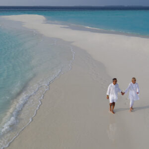 A couple dressed in white walks hand in hand along a sandy beach with turquoise waters on both sides, reminiscent of a Maldives honeymoon resort. The beach stretches into the distance, forming a narrow strip of land surrounded by clear blue ocean. The sky is overcast with light clouds.