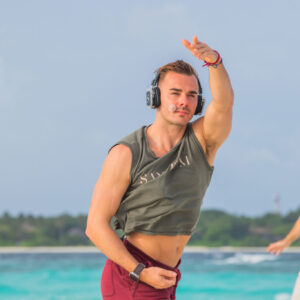 Mel Douglas, wearing headphones, a grey sleeveless shirt, and red shorts, dances energetically on a sandy beach at Soneva. The sky is overcast, with the ocean providing a serene backdrop.