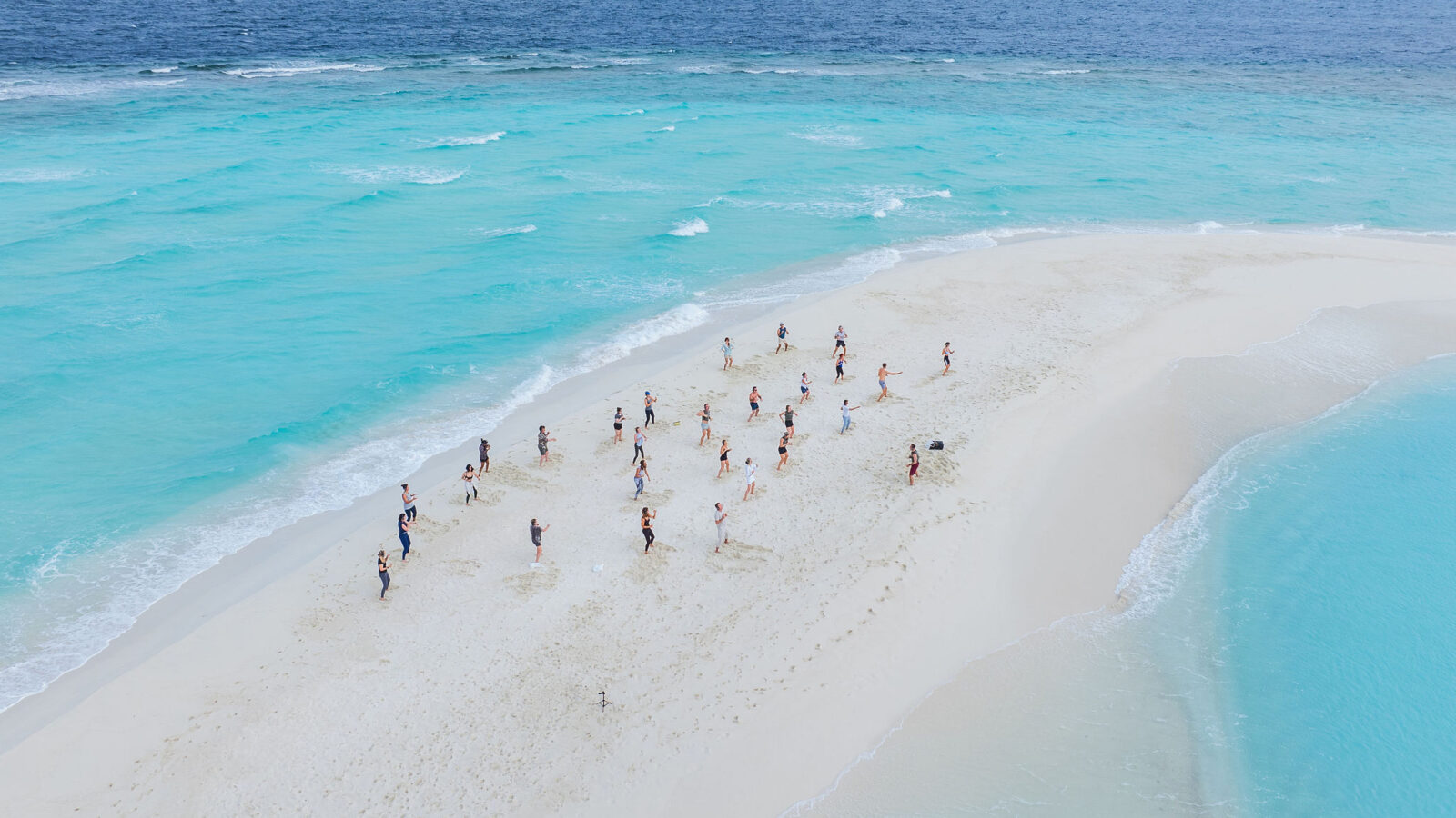 Aerial view of a sandbar at Soneva, surrounded by turquoise water. A group of people, including Mel Douglas, is scattered along the sandbar enjoying the sunny weather. Waves gently lap at the shoreline, creating a striking contrast between the white sand and deep blue ocean.