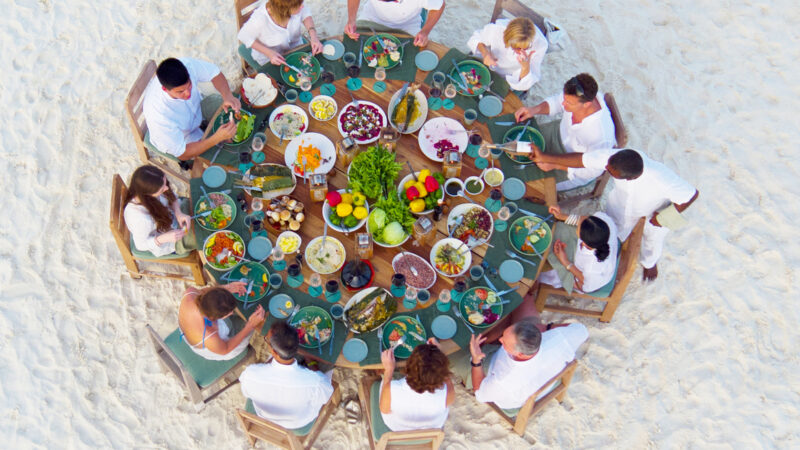 Aerial view of a group of people seated in a circle around a large table on a sandy beach. The table, resembling a Soneva Award festivity, is filled with various colorful foods and dishes. The people are dressed in white, creating a striking contrast with the sand and vibrant food.