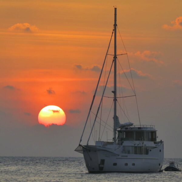 A luxurious white sailboat is anchored on the calm waters of the Maldives during a vibrant sunset. The sun, partially obscured by light clouds, casts an orange and pink glow across the sky and reflects on the water's surface, epitomizing a luxury yacht holiday at Soneva.