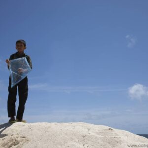A boy stands on a sandy hill in the Maldives, holding a transparent kite against a clear blue sky. The background shows a serene sky with a few scattered clouds. The overall atmosphere is calm and bright, capturing the Namoona spirit of simplicity and beauty.