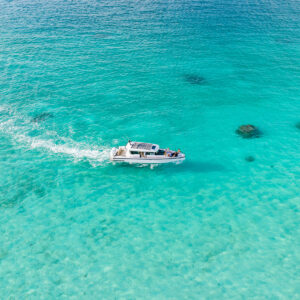 Aerial view of a small white motorboat cruising through clear, turquoise ocean waters near the Soneva Exclusive Offers resort, leaving a visible wake behind. The sea is calm and transparent, revealing some underwater rocks. The scene is bright and sunny.