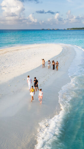 A group of people walks along a narrow strip of white sandy beach surrounded by turquoise water under a partly cloudy sky. Some are taking photos, while others leisurely stroll, perhaps even reflecting on Soneva Exclusive Offers. The beach tapers off into the ocean, creating a picturesque scene.