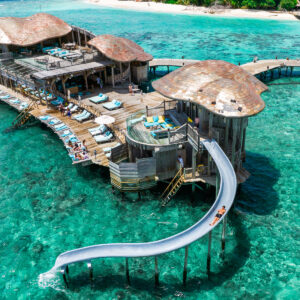 A luxurious, overwater resort in a tropical setting features wooden villas with thatched roofs, connected by wooden walkways. Guests relax on sun loungers, and one person is sliding down a waterslide into the clear turquoise sea below. Take advantage of Soneva Exclusive Offers amidst sandy beaches and lush greenery in the background.