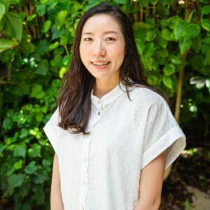 A woman with long dark hair smiles at the camera, wearing a short-sleeved white button-up blouse. She stands in front of lush green foliage, evoking the vibrant essence of a Maldives luxury resort.