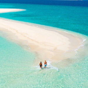 A couple walks along a sandbar in clear turquoise waters, surrounded by the deep blue sea under a clear sky. An island is visible in the distance on the horizon.