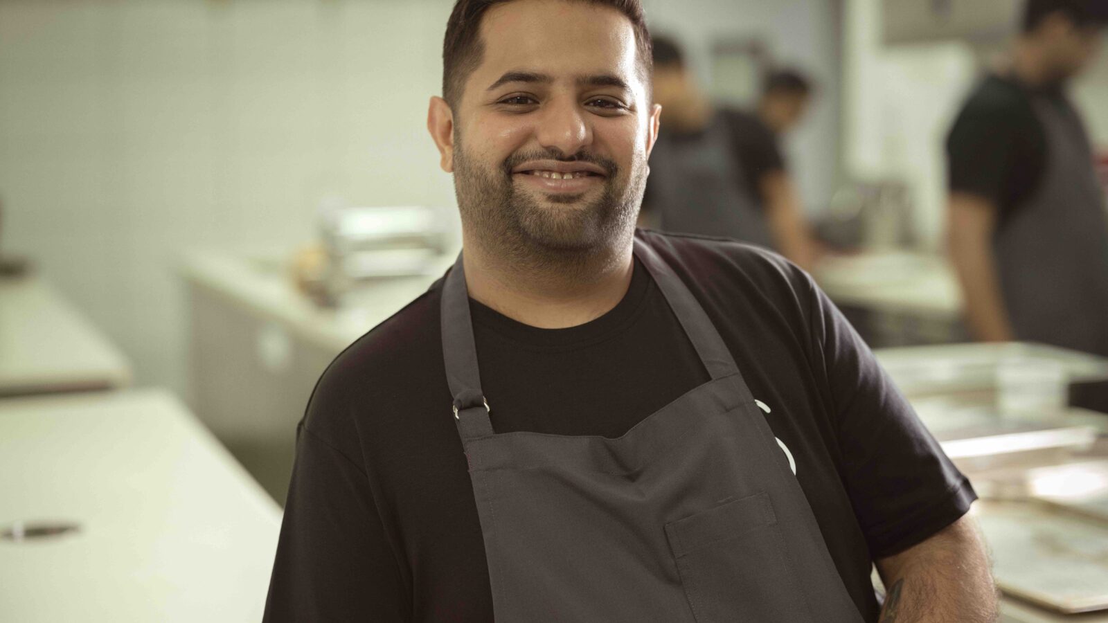 A man wearing a black shirt and a gray apron stands smiling in a bright kitchen. The blurred background shows a counter and other people working.