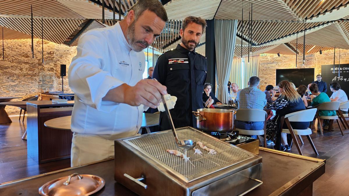 Two chefs cooking at a metal grill in a modern restaurant with wooden ceiling panels. One chef is placing food on the grill, while the other watches. A group of people is seated in the background, observing the demonstration.