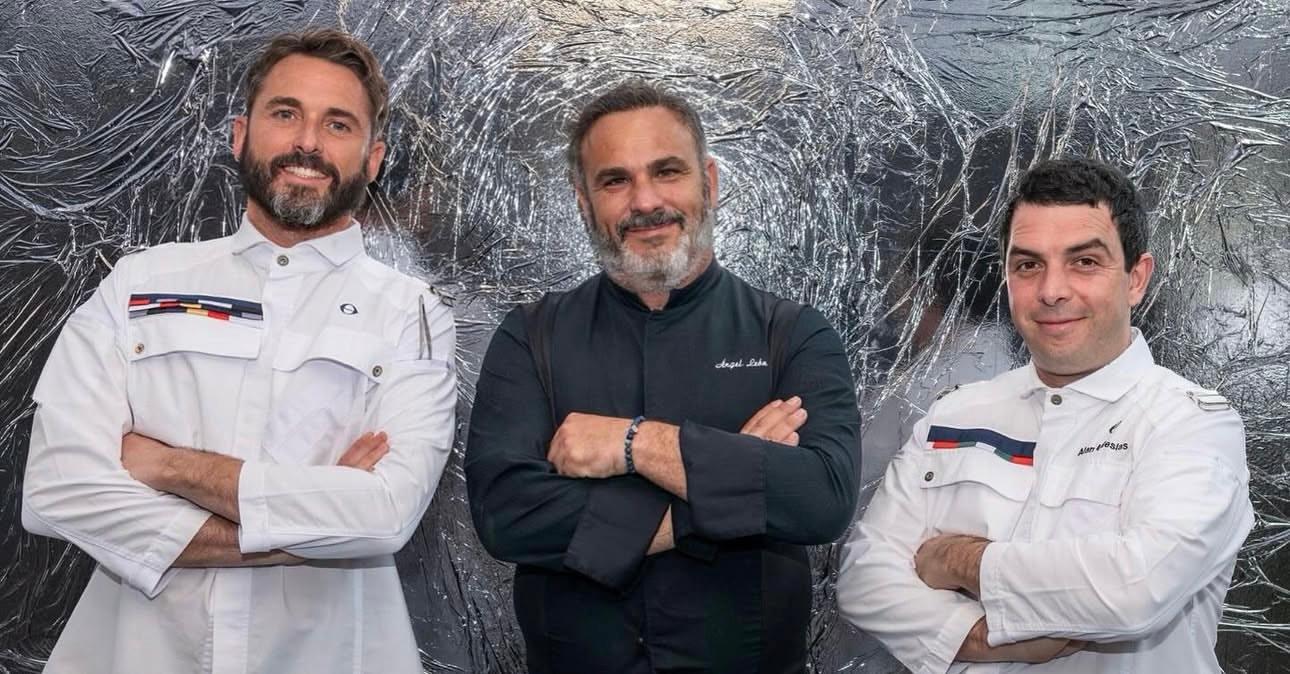 Three chefs stand with arms crossed, posing in front of a shiny, crinkled metallic backdrop. Two wear white uniforms with colorful trim while the one in the center wears a dark uniform. They are all smiling softly.