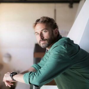 Man with a beard and green shirt leans on a railing, looking thoughtfully into the distance. He is indoors, and the background is softly lit with large windows and hanging lights, giving an airy, spacious feel to the room.