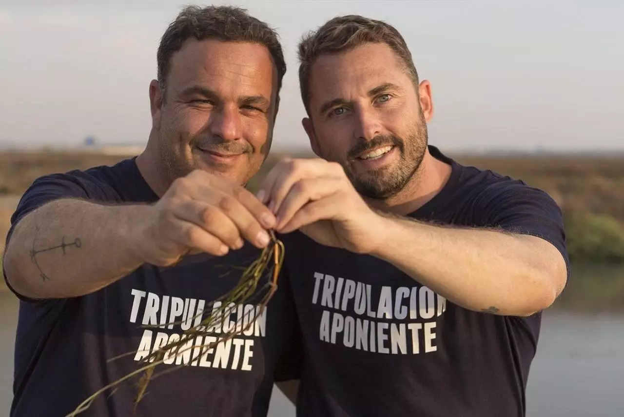 Two smiling individuals in navy shirts with &quot;TRIPULACIÓN APONIENTE&quot; printed on them, standing outdoors. They hold a small plant together, with a natural landscape in the background.