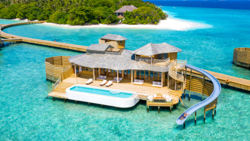 A luxurious overwater villa at Soneva Fushi Villas in the Maldives sits on turquoise ocean water, featuring a private pool, multiple decks with loungers, and a waterslide extending from a balcony. Wooden walkways connect the villa to a lush tropical island with white sandy beaches in the background.