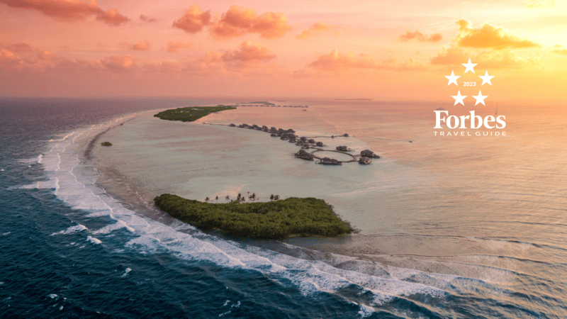 A stunning aerial view of the Maldives at sunset, featuring overwater bungalows at the luxurious Soneva resort stretching along the shallow turquoise water. The ocean waves gently lap against the shore. The sky is painted with hues of orange and pink. The Forbes Travel Guide logo is visible.