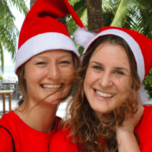 Two smiling women wearing red Santa hats and red shirts stand close together, with tropical palm trees and a beach in the background. They seem to be enjoying a festive Family Holiday in the Maldives, savoring a joyful moment at Soneva's luxurious resort.