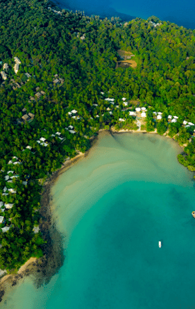 An aerial view of a lush green peninsula extending into a clear blue sea, showcasing Thailand's exclusive experience. The peninsula boasts scattered white buildings among dense trees, a curved sandy beach with a small pier stretching into the water, and the surrounding ocean displaying various shades of blue.