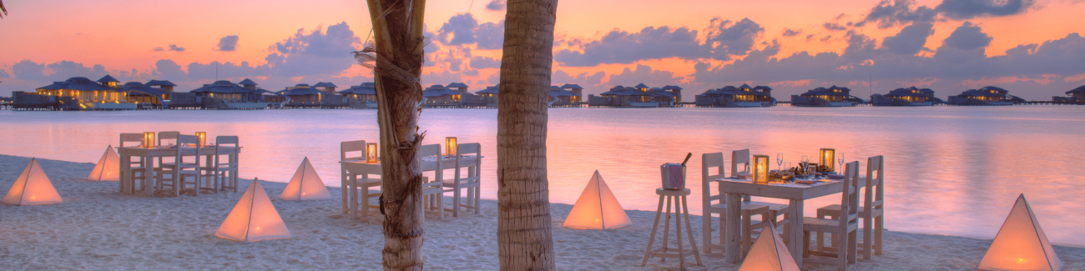 A picturesque beach setting at sunset with dining tables set up on the sand, surrounded by cozy pyramid lights. The calm ocean and bungalows in the background enhance the serene and romantic ambiance, epitomizing Soneva Maldives' luxury experience.