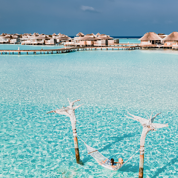 A person relaxes in a hammock suspended over crystal-clear turquoise waters, supported by two wooden poles, with overwater bungalows and a vibrant blue sky visible in the background—a quintessential Soneva Maldives holiday.