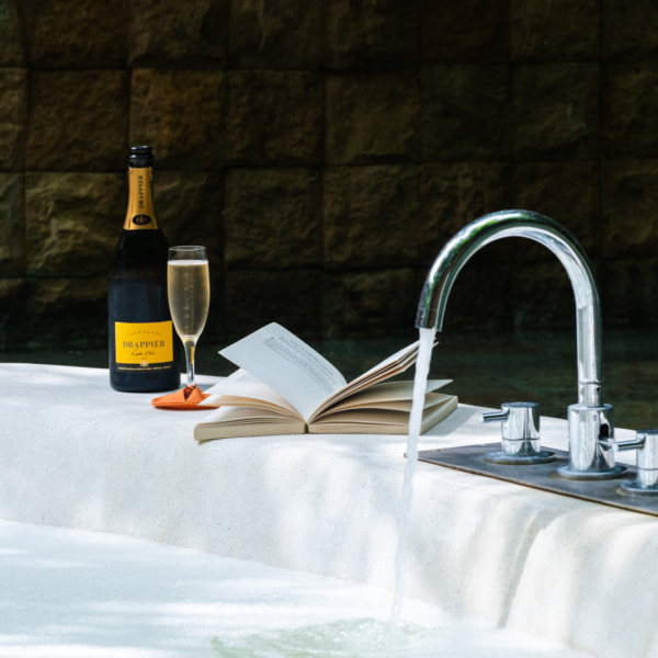 A luxurious bathtub with water flowing from a sleek silver faucet. Beside the tub is an open book, a glass of champagne, and a bottle of Drappier champagne. The background features a stone wall, enhancing the serene and opulent atmosphere reminiscent of celebrating Chinese New Year at Soneva in the Maldives.