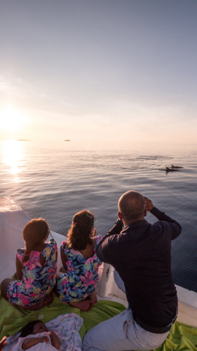 A family of four enjoys a peaceful moment on a boat at sunset, part of the Maldives Exclusive Experience with Soneva. An adult points excitedly at a dolphin swimming nearby while two children in floral dresses watch. Another adult lies nearby, partially covered by a green blanket. The calm sea reflects the warm hues of the setting sun.