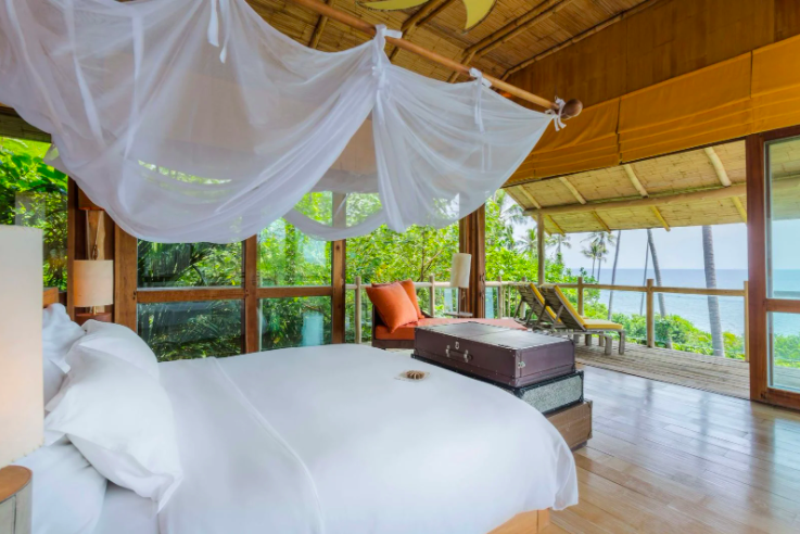 A well-lit bedroom with a large white bed under a mosquito net canopy. Wide windows provide a view of lush greenery and the ocean, perfect for a Thailand family holiday. A wooden trunk sits at the foot of the bed, and a spacious balcony with lounge chairs offers additional views, reminiscent of Soneva's elegance.