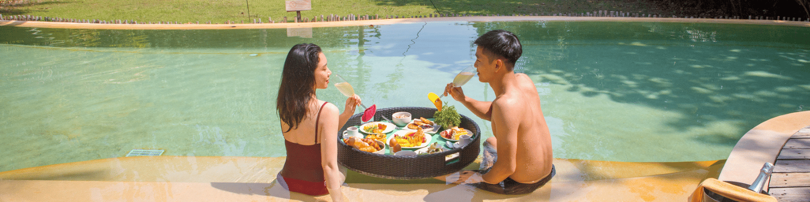 A woman and a man enjoy a meal on a floating tray in a swimming pool at Soneva, Thailand. The tray is filled with various dishes and drinks. Seated on the edge of the pool, they smile and converse, surrounded by lush greenery with sunlight casting a warm glow on their exclusive experience.