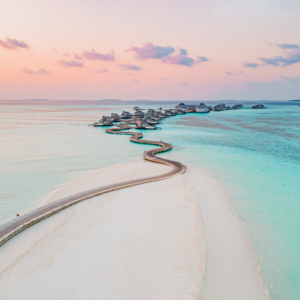 Aerial view of a winding boardwalk leading to overwater villas set against a serene turquoise ocean and pink-hued sunset sky. The scene, enhanced by Soneva Exclusive Offers, features white sandy beaches and calm, clear water, creating a tranquil and picturesque tropical paradise.