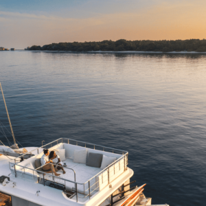 A person sits on the upper deck of a sailboat, reading a book and enjoying the serene surroundings during sunset. The boat is on calm waters near a tree-lined shore under a clear sky, as if part of Soneva Exclusive Offers designed for ultimate tranquility.