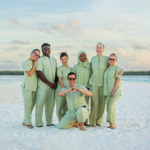 A group of seven people, dressed in matching light green outfits, pose together on a white sandy beach at a Maldives luxury resort. One person kneels in the front making a heart shape with their hands, while the others stand behind, smiling and hugging each other. The background features a calm sea and pastel sky.