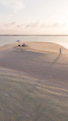 A serene beach scene with a tiny sandbar surrounded by calm waters, offering a true Maldives exclusive experience. Two people sit under a white umbrella, while a third person walks on the sand, casting a long shadow in the sunlight. The sky is partly cloudy, and the horizon is visible in the background.