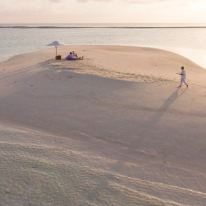 A serene beach scene with a tiny sandbar surrounded by calm waters, offering a true Maldives exclusive experience. Two people sit under a white umbrella, while a third person walks on the sand, casting a long shadow in the sunlight. The sky is partly cloudy, and the horizon is visible in the background.