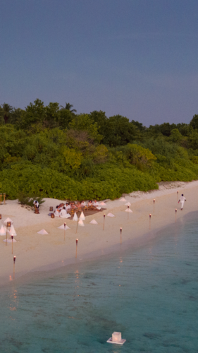 A serene beach scene at sunset, featuring a small wedding ceremony on the sandy shore. Guests are seated, and the aisle is lined with tiki torches leading to an altar near the water's edge. Dense greenery and palm trees form a backdrop in this Maldives Exclusive Experience courtesy of Soneva.