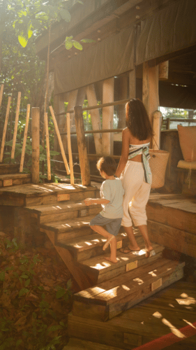 A woman and a young child walk up wooden steps towards a rustic, treehouse-like structure surrounded by lush greenery. Sunlight filters through the trees, creating a warm and inviting atmosphere. On this family holiday at Soneva in the Maldives, the woman holds the child's hand, guiding them up the stairs.