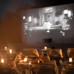 People sitting in wooden chairs with small candlelit tables, watching a black-and-white movie projected onto a large outdoor screen at night. The scene is cozy and surrounded by a warm ambiance, reminiscent of Maldives dining experiences at Soneva Fushi.