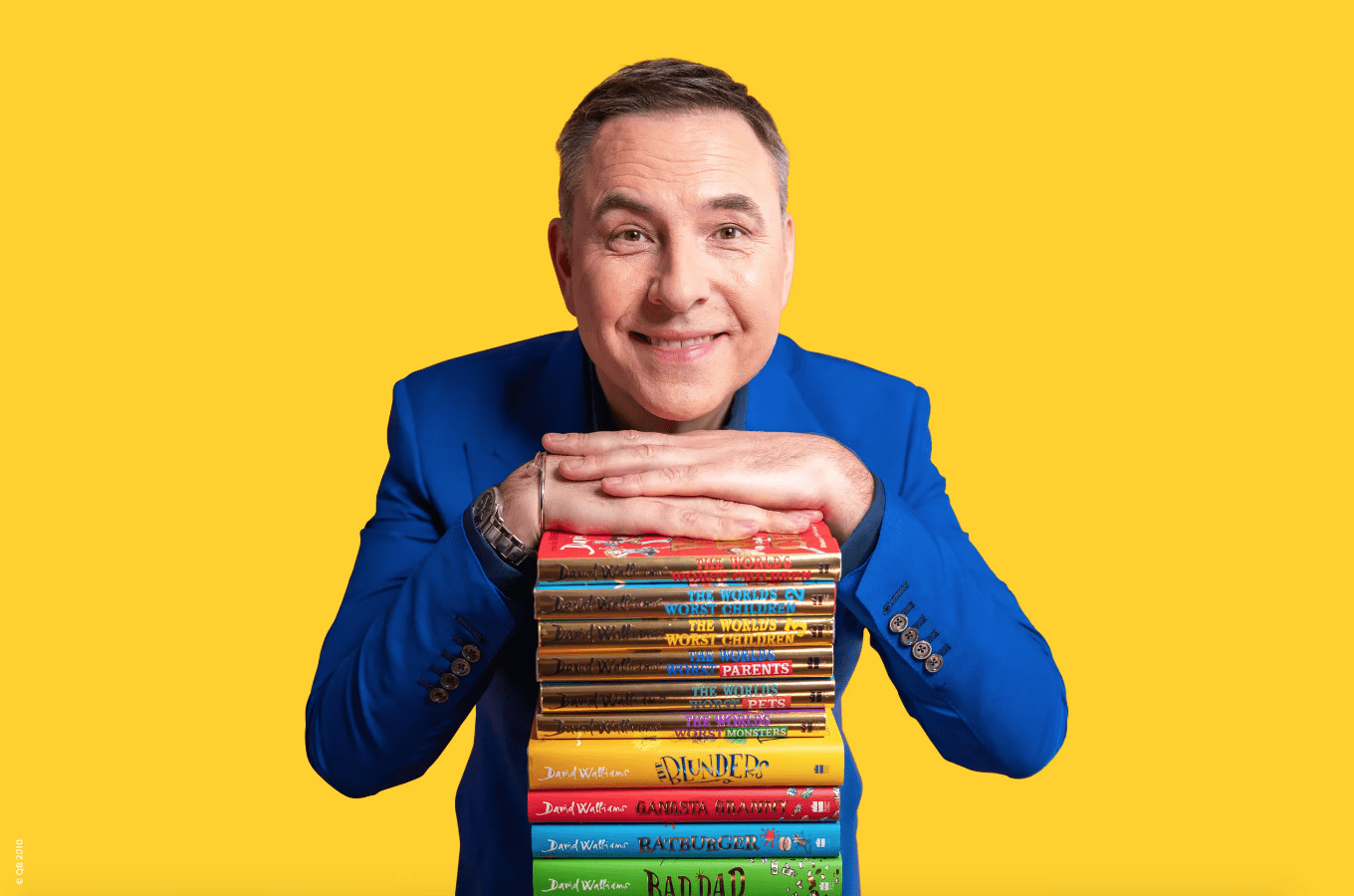 A smiling man in a blue jacket rests his hands on a stack of colorful books against a bright yellow background. The books have playful, illustrated covers.