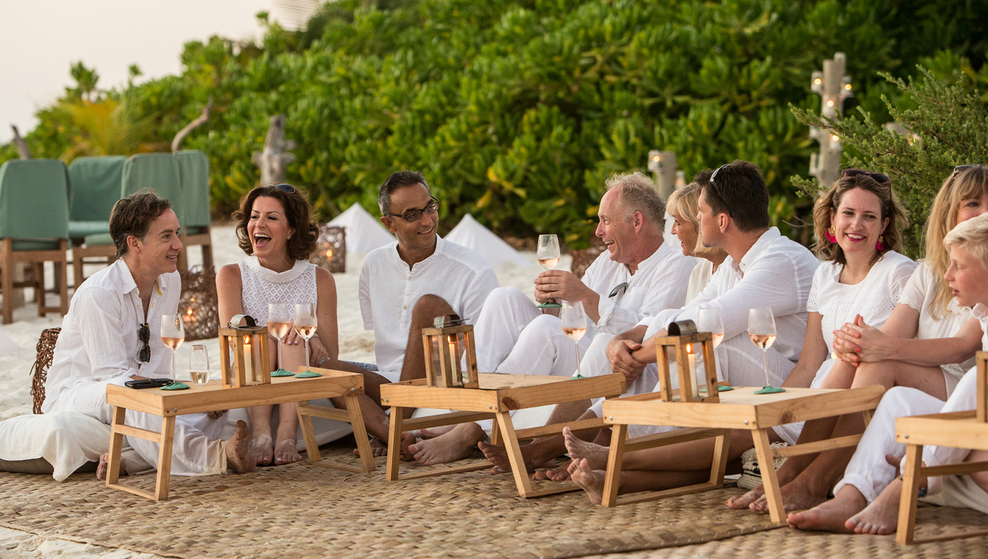 A group of people dressed in white sit on a sandy beach, gathered around low wooden tables. They are smiling, talking, and holding drinks. Lanterns adorn the tables, enhancing the relaxed ambiance with lush greenery in the background. The scene exudes joy akin to Soneva Exclusive Offers' promise of blissful experiences.