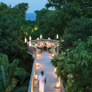 A group of people dine on a round, elevated wooden deck surrounded by lush, green trees. It is evening, and the path to the deck is illuminated by soft, warm lights. A person is walking towards the deck along the lit pathway at Soneva Fushi in the Maldives, with the ocean visible in the background.